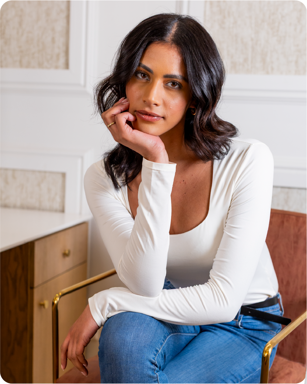 Beatufiul woman in a white long-sleeve top and blue jeans sitting indoors. Her hair is shinny and styled to perfection. 