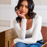 Woman in a white long-sleeve top and blue jeans sitting indoors.