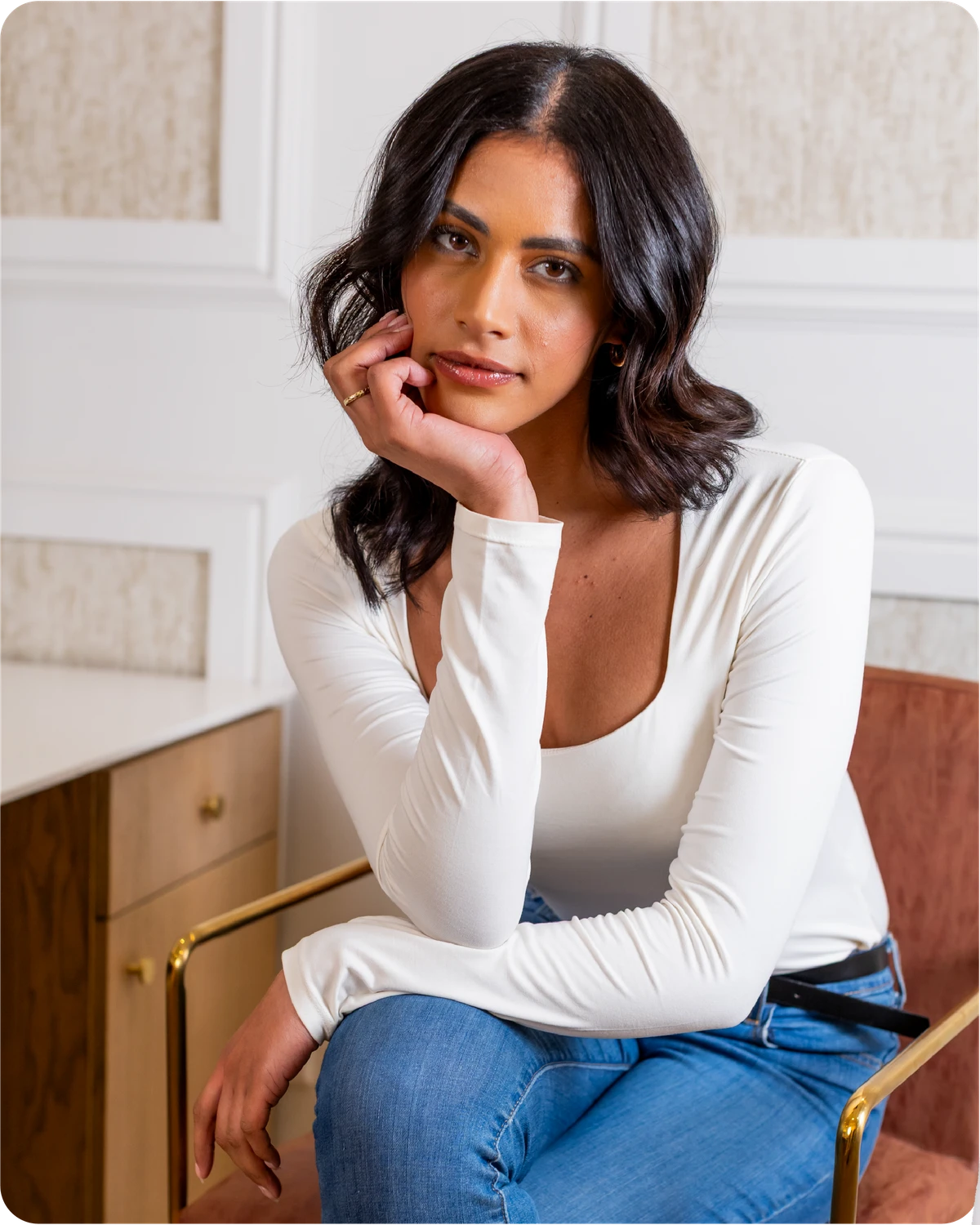 Woman in a white long-sleeve top and blue jeans sitting indoors.
