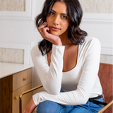 Woman in a white long-sleeve top and blue jeans sitting indoors.