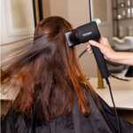 Moyoko Aria E9 is a lightweight, high-speed hairdryer that delivers salon-level results. Image shows a lady using a black hairdryer to dry their hair in a salon setting