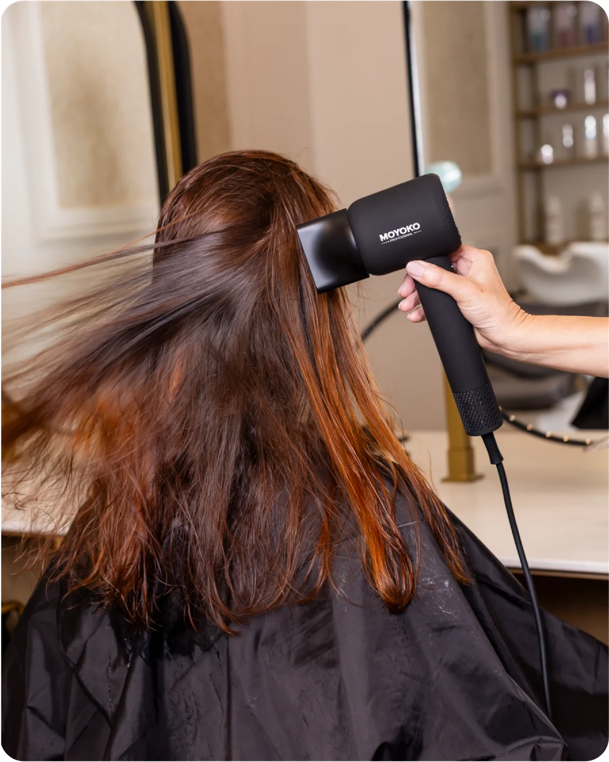 Moyoko Aria E9 is a lightweight, high-speed hairdryer that delivers salon-level results. Image shows a lady using a black hairdryer to dry their hair in a salon setting
