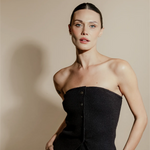Woman wearing a black strapless top against a beige background. Her hair is perfectly pulled back and no flyways as she uses Sleek back hair wax stick